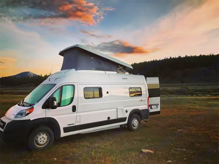 Sunset views from your rv rental! This white pop-top camper van is parked in a vast field with mountains and trees.. Winnebago Solis 2021