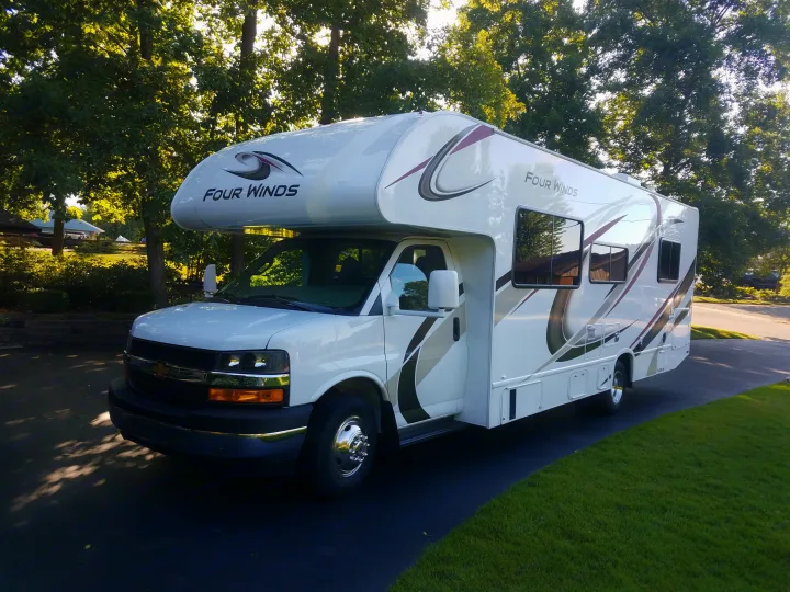 White Four Winds Class C RV rental parked on a driveway, surrounded by green trees and grass.. Thor Four Winds 2020