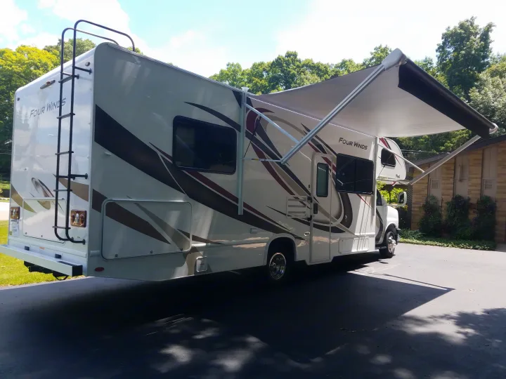 This Four Winds motorhome, perfect for an RV rental, is parked with its awning extended, showing the side and rear ladder.. Thor Four Winds 2020