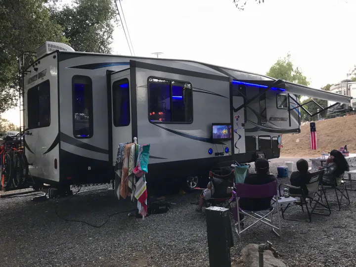 An 'Arctic Wolf' RV rental with blue lights is parked at a campsite. People relax in chairs watching outdoor TV.. Forest River Cherokee Wolf Pack 2019