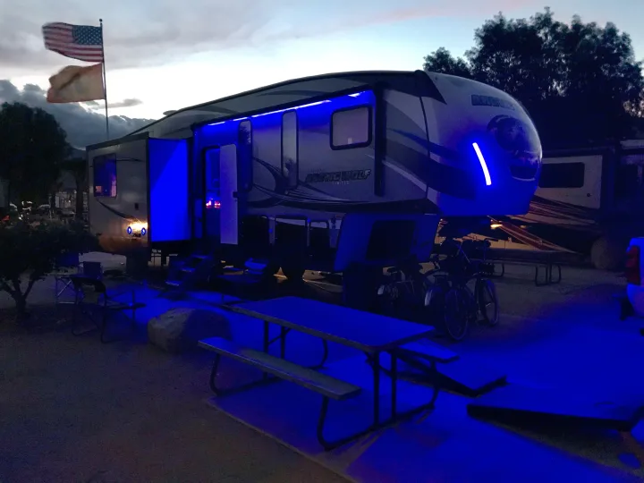 RV rental: A large RV shines with vibrant blue lights at a campsite with flags, bikes, and a picnic table at dusk.. Forest River Cherokee Wolf Pack 2019