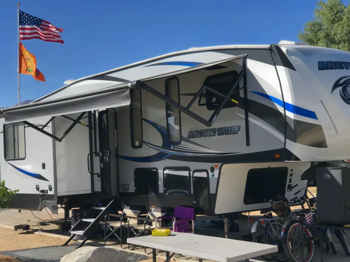This Cherokee Arctic Wolf fifth-wheel RV, possibly an RV rental, is set up at a sunny campground with its awning extended, flags flying, and picnic area ready.. Forest River Cherokee Wolf Pack 2019