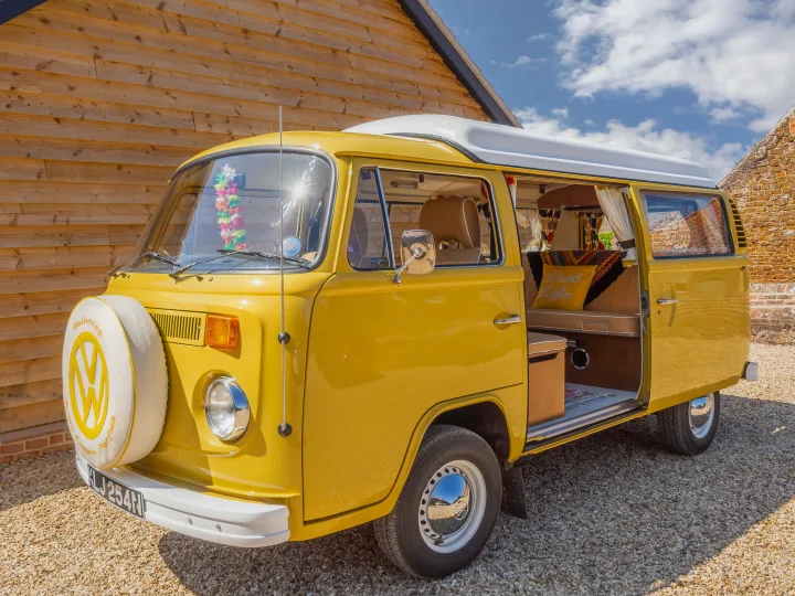 Bright yellow vintage VW campervan, side door open, perfect rv rental for adventure. Parked on gravel by a wooden barn.. Volkswagen 2.0L T2 Late Bay 1975