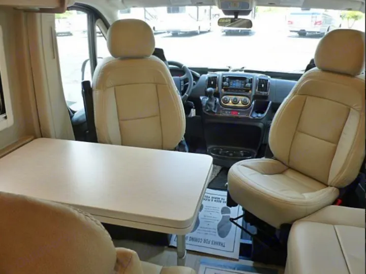 RV rental interior: Light beige front seats, dashboard, and a dining table are visible, showing a spacious living area.. Winnebago Travato 2020