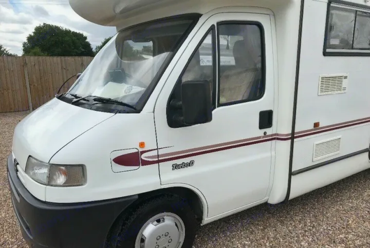 White motorhome (RV) for rv rental. Front-left view shows driver's cab, striped side, and vents on gravel.. Peugeot Boxer Elddis Auto Stratus EB 1999