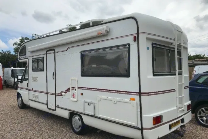 White RV rental, a recreational vehicle, parked on gravel. Side view shows an awning, windows, and rear ladder.. Peugeot Boxer Elddis Auto Stratus EB 1999