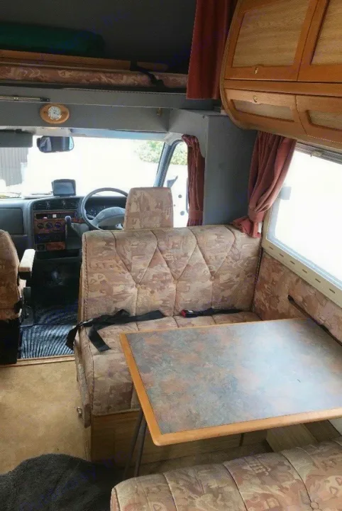 This RV rental's interior shows a dining area with patterned seats, seatbelts, an overhead bunk, and front cockpit.. Peugeot Boxer Elddis Auto Stratus EB 1999