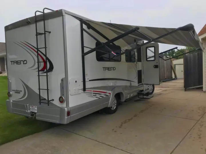 White "TREND" RV rental parked on a driveway with awning extended, door open, and rear ladder visible.. Winnebago Trend 2017
