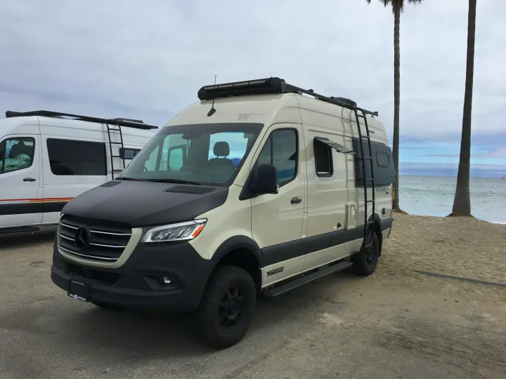 This Winnebago Revel rv rental, a beige Mercedes Sprinter van, is parked on a sandy path by the ocean.. Winnebago Revel 2020