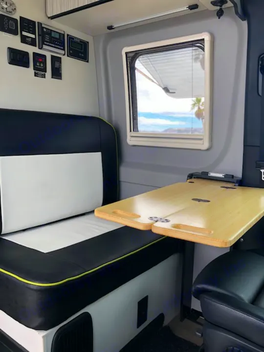 Inside a modern RV rental: comfortable black/white seating, wooden foldable table, window showing an outdoor view, and control panels.. Winnebago Revel 2020