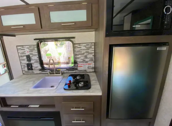 RV rental's compact kitchen with stainless fridge, microwave, sink, 2-burner stovetop, and tiled backsplash.. Dutchmen Coleman 2021