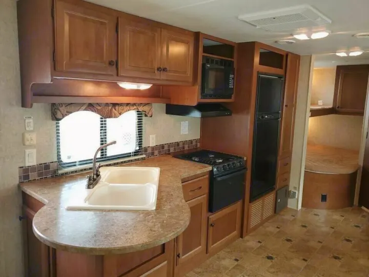RV rental interior: compact kitchen with wood cabinets, sink, stove, microwave, and fridge. A sleeping area is in the back.. Heartland Prowler 2012