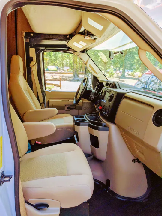 Front cabin of an RV rental with comfy seating and dashboard, offering a clear view of a forest campground.. Nexus RV Triumph 2020