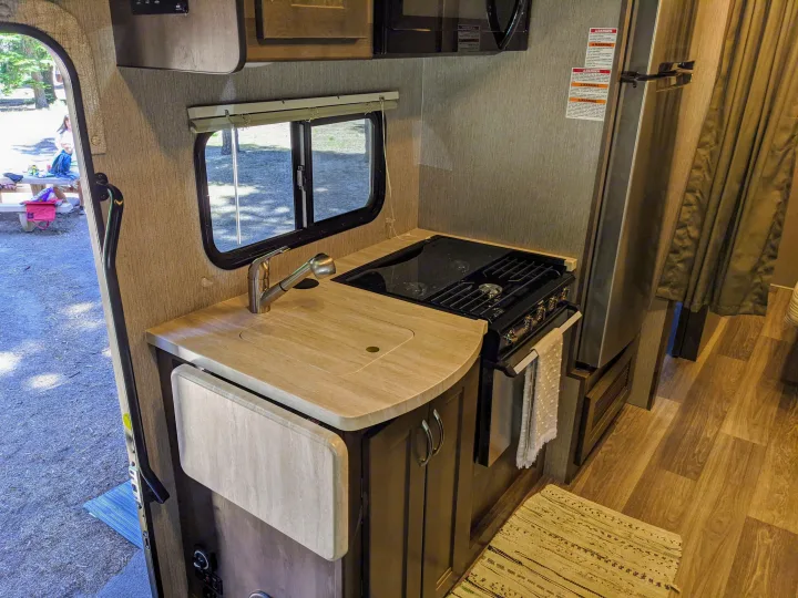 Inside an RV rental: Compact kitchen with sink, stove, microwave, and fridge. View outside to campground.. Nexus RV Triumph 2020