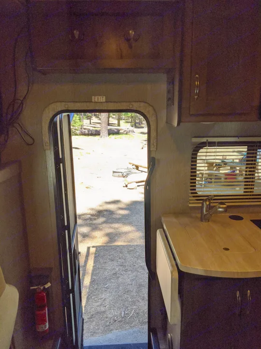 RV rental interior view: open door to a sunny campsite, featuring kitchen counter and fire extinguisher.. Nexus RV Triumph 2020