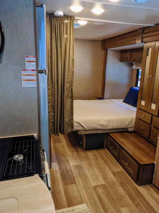 Inside an RV rental, a comfy bedroom with privacy curtain and clever storage is visible, plus a peek at the kitchen stove.. Nexus RV Triumph 2020