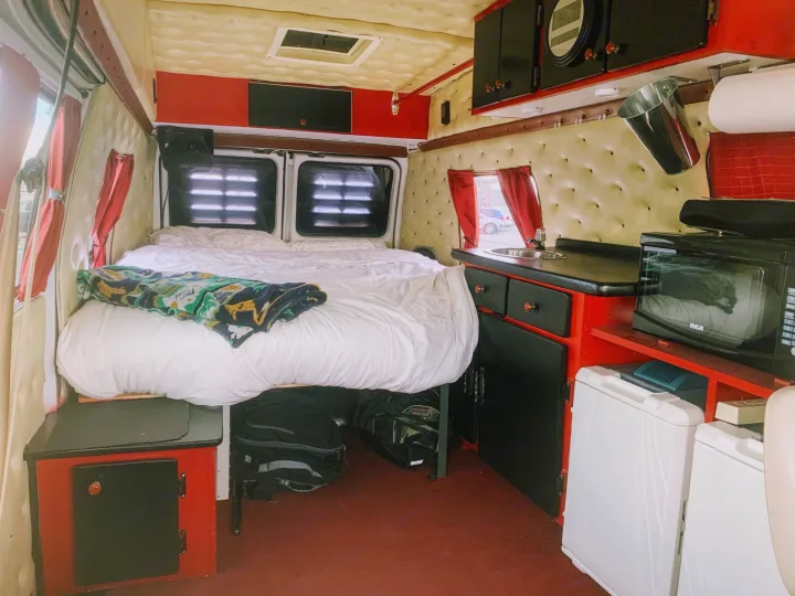 Explore this cozy campervan RV rental interior featuring a large bed, compact kitchen with sink, microwave, and fridge, all with stylish red and black accents.. Chevrolet G20 1983