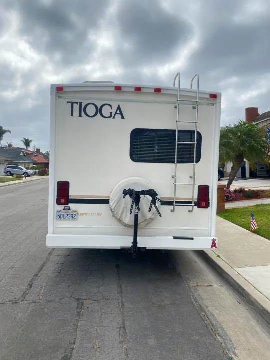 Rear view of a white Tioga Fleetwood RV rental, featuring a bike rack, ladder, and spare tire, parked on a street. Ready for travel!. Fleetwood Tioga 2003
