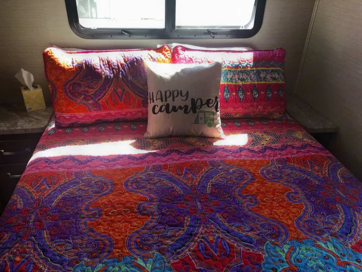 RV rental's cozy bedroom features vibrant bedding & a "HAPPY camper" pillow, bathed in sunlight.. Jayco Greyhawk 2019