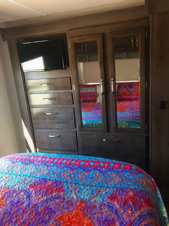 This RV rental bedroom features a TV, drawers, mirrored wardrobe, and a vibrant, patterned quilt, offering cozy travel comfort.. Jayco Greyhawk 2019