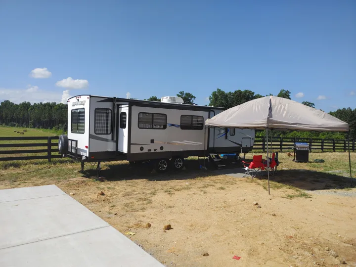 RV rental camper with slide-out, canopy, grill & chairs at a sunny, rural campsite by a field and forest. Relaxing outdoor stay.. Forest River Cherokee Alpha Wolf 2021