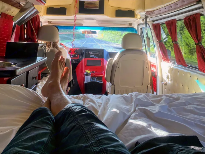 Feet up on the bed inside an rv rental, cruising down a green highway. Perfect road trip comfort in this camper van.. Chevrolet G20 1983