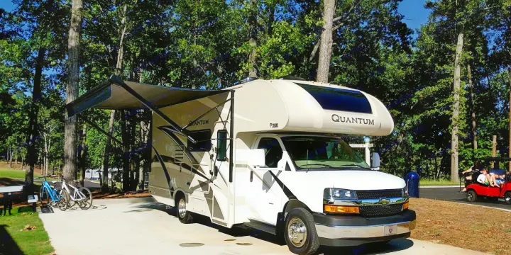 A Thor Quantum rv rental is parked with its awning out at a wooded campsite, featuring bikes and a golf cart nearby.. Thor Quantum 2020