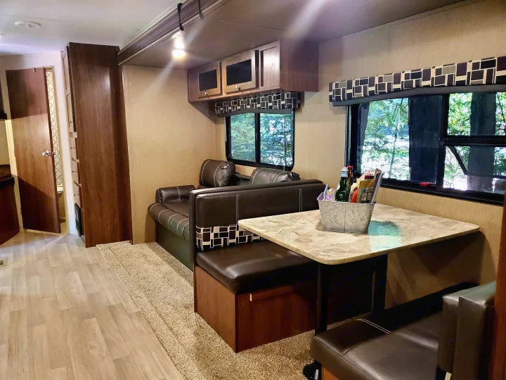 Inside an RV rental: a bright, comfortable living space with a dining booth, couch, and large windows revealing green nature.. Coleman 285bh 2017