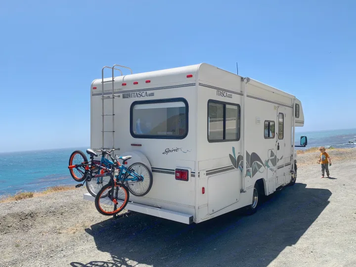 An RV rental parked on a sunny coast with bikes attached, overlooking the blue ocean.. Itasca Spirit 1999