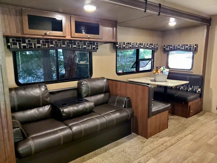 An rv rental's interior showing a cozy brown leather sofa with cupholders, a dinette, and bright windows.. Coleman 285bh 2017