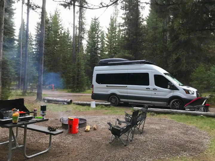 RV rental camper van at a smoky forest campsite. Picnic table cooking, active campfire, and chairs set up for relaxation.. Ford Transit Custom 2015