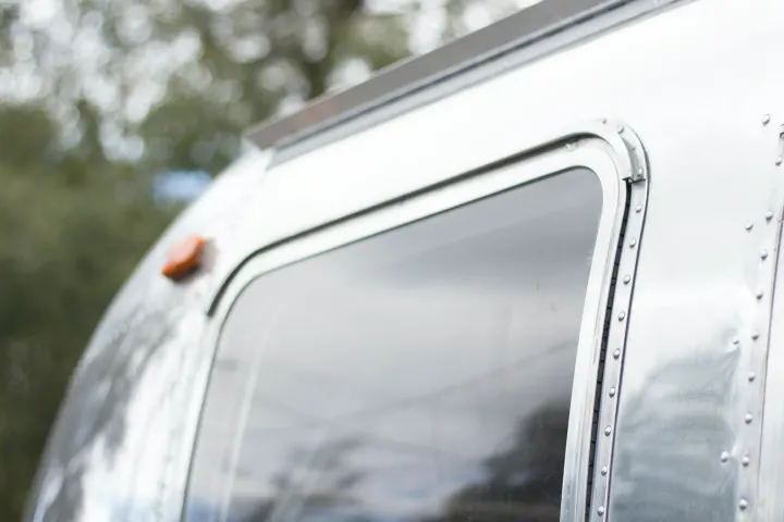 Curved side of a classic silver RV rental, highlighting its window and riveted aluminum construction against blurred nature.. Airstream Bambi 2021