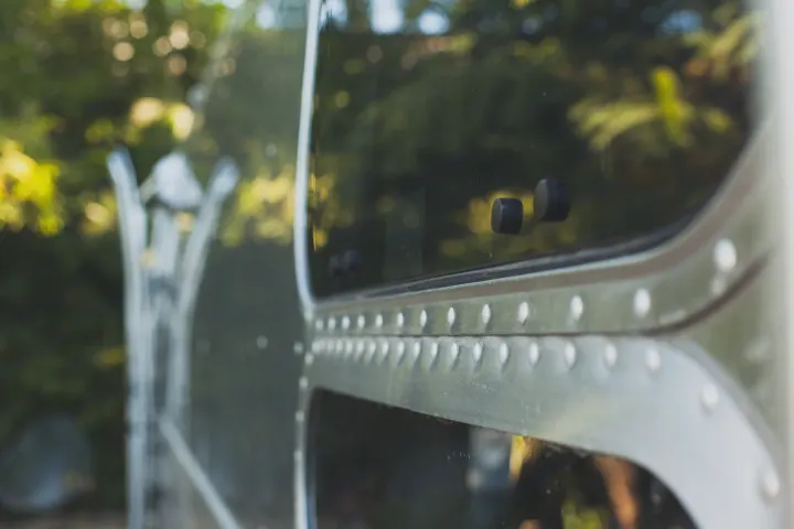 Close-up of a vintage-style RV rental's riveted aluminum side, featuring a window reflecting green, blurred foliage.. Airstream Bambi 2021