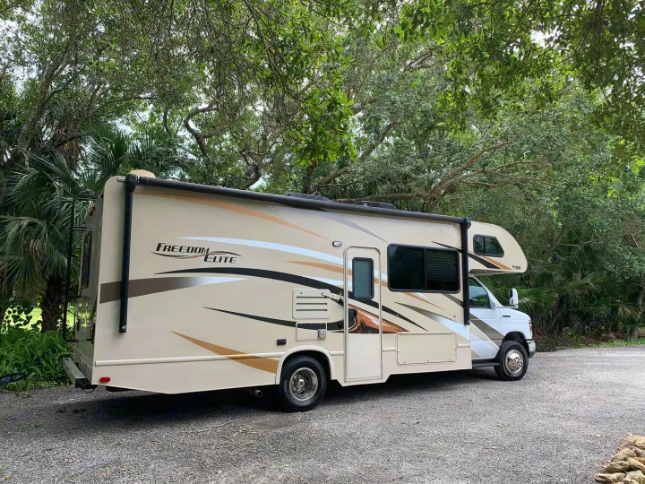 A Freedom Elite Class C RV rental is parked on a gravel lot, nestled among green trees, perfect for your next travel adventure.. Thor Motor Coach Freedom Elite 2017
