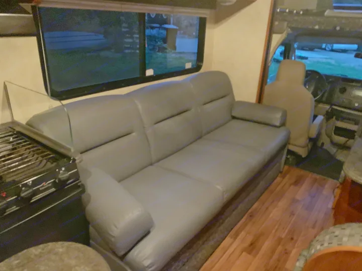 Spacious RV rental interior with a long gray sofa, kitchen stove, and large window overlooking a dark exterior.. Coachmen Freelander 2012