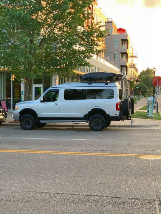 Silver custom off-road van, an ideal RV rental, with roof cargo box & spare tire, parked on a city street.. Roadtrek N6-Active 2013