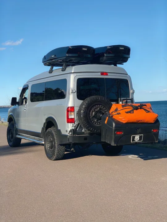 A modified silver RV rental, equipped with rooftop cargo boxes and rear adventure gear, parked by a vast blue lake.. Roadtrek N6-Active 2013