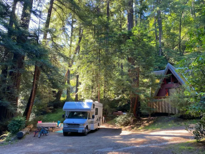 A white RV rental is parked at a spacious forested campsite, featuring a picnic table, bikes, and a nearby A-frame cabin.. Itasca Spirit 1999