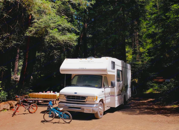 White RV rental parked at a shady forest campground with bikes and a picnic table.. Itasca Spirit 1999
