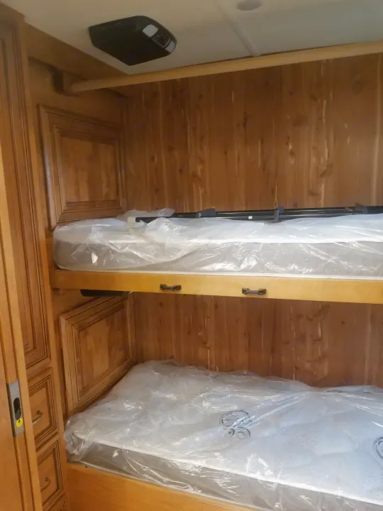 RV rental interior features two new bunk beds with mattresses in plastic, wood paneling, and an entertainment system.. American Coach 42B 2019