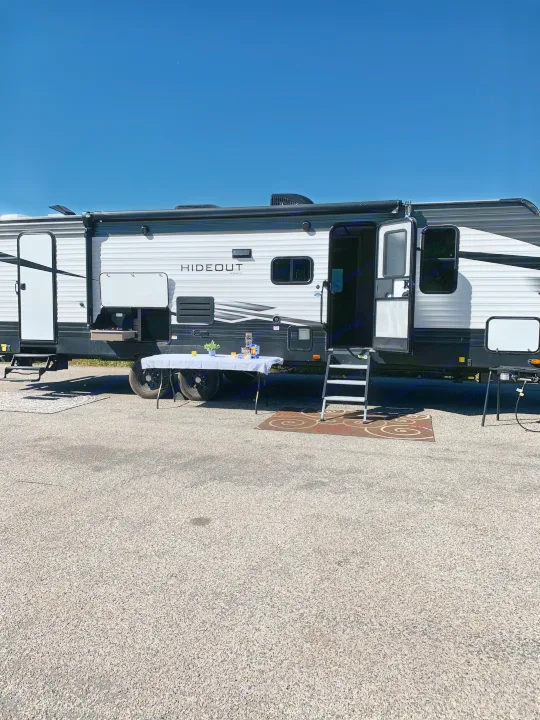 This RV rental is set up at a sunny campsite with an extended awning, outdoor kitchen, and a table on gravel.. Keystone Bunkhouse 2020