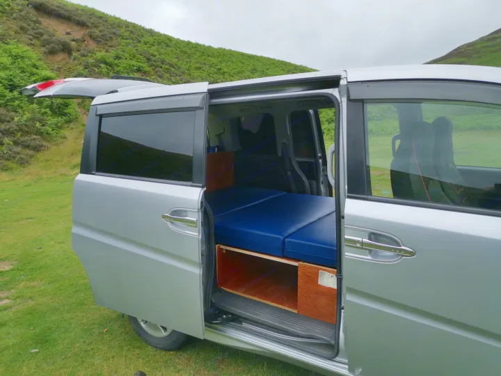Explore the outdoors! This customized silver camper van, ideal for an RV rental, reveals its blue bed and storage with doors open.. honda stepwagon 2006
