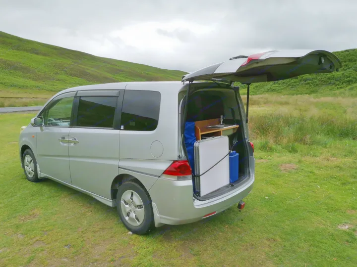 Silver camper van with open trunk showing a portable kitchen setup, parked on green grass near hills. Perfect for rv rental camping adventures!. honda stepwagon 2006