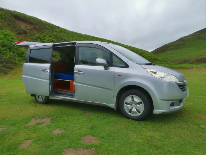 Silver converted minivan, ideal for rv rental, parked on grassy hills with doors open, showing a cozy bed setup.. honda stepwagon 2006