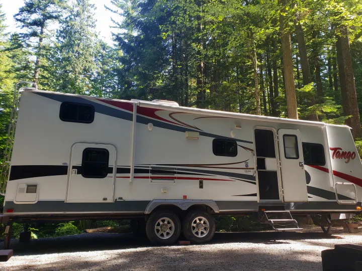 Experience nature with this Tango rv rental, a white travel trailer with red & grey accents, parked in a lush forest campground.. Pacific Coachworks Tango 2010