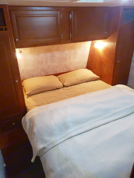 Cozy bed with light sheets and white blanket in an RV rental, featuring overhead cabinets, reading lights, and an outlet.. Pacific Coachworks Tango 2010