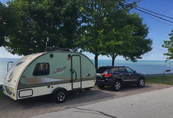 Light green R-Pod camper (an rv rental) hitched to a dark Jeep, parked roadside by a serene blue lake with lush green trees.. Other Other 2016