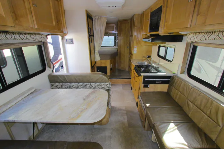 Interior of an rv rental with a dinette, functional kitchen (stove, sink, microwave), and overhead cabinets.. Coachmen Leprechaun 2020