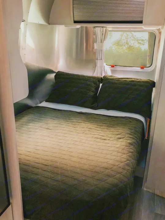Inside a comfortable RV rental: A cozy bedroom with a quilted bed, dark green pillows, and a window showing forest views.. Airstream Bambi 2021