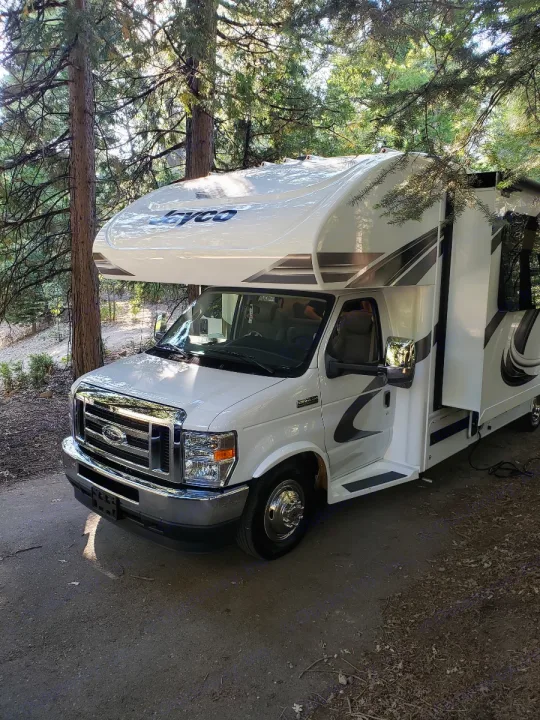 White Jayco Class C RV rental parked under tall trees on a sunny day. A perfect getaway for camping and outdoor adventure.. Jayco Greyhawk 2021